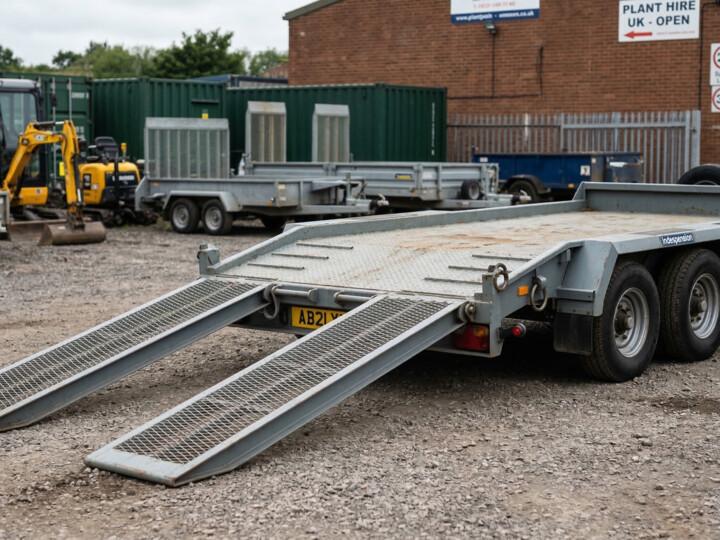 Heavy duty plant trailer with twin loading ramps extended at a UK hire yard with mini-digger in background